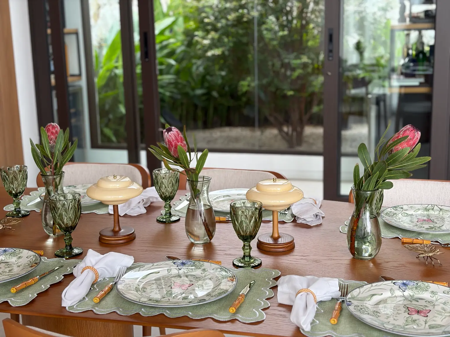 Mesa posta com proteas rosas, porcelana e taças de vidro verde-oliva